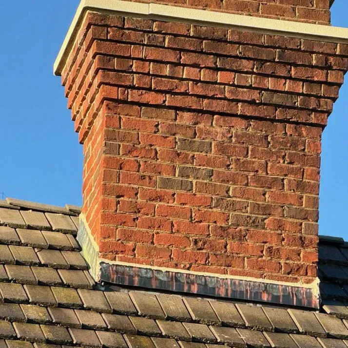 Close-up of a brick chimney on a roof with shingles and a clear blue sky in the background.