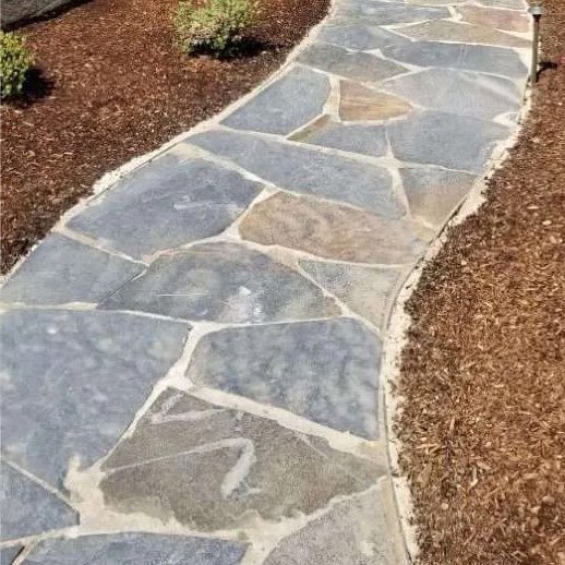 A curved stone pathway with irregularly shaped stones and light-colored mortar, bordered by mulch and shrubbery.