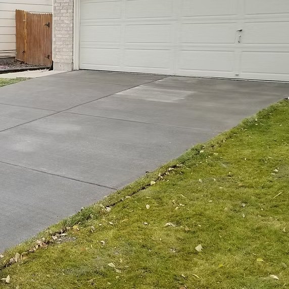 Concrete driveway next to a grassy yard with some scattered leaves in front of a white garage door and a wooden gate.