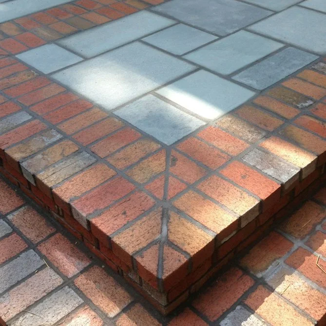 Close-up of a brick sidewalk corner with a tiled stone pattern in the center, showing the contrast between red bricks and gray stones.