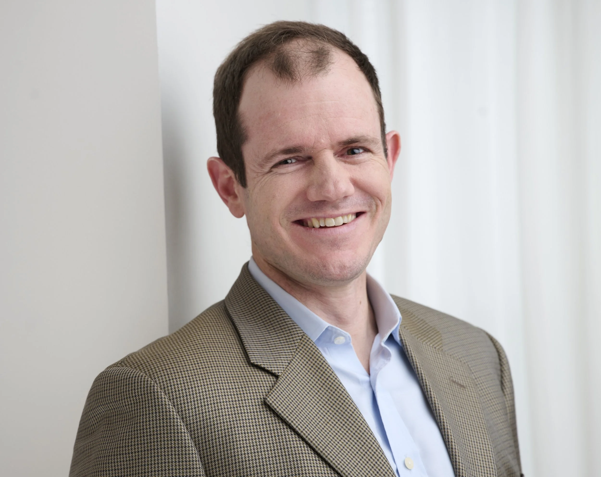 Close-up of a smiling man with short brown hair, wearing a beige checkered blazer and a blue collared shirt, standing indoors against a gray wall.