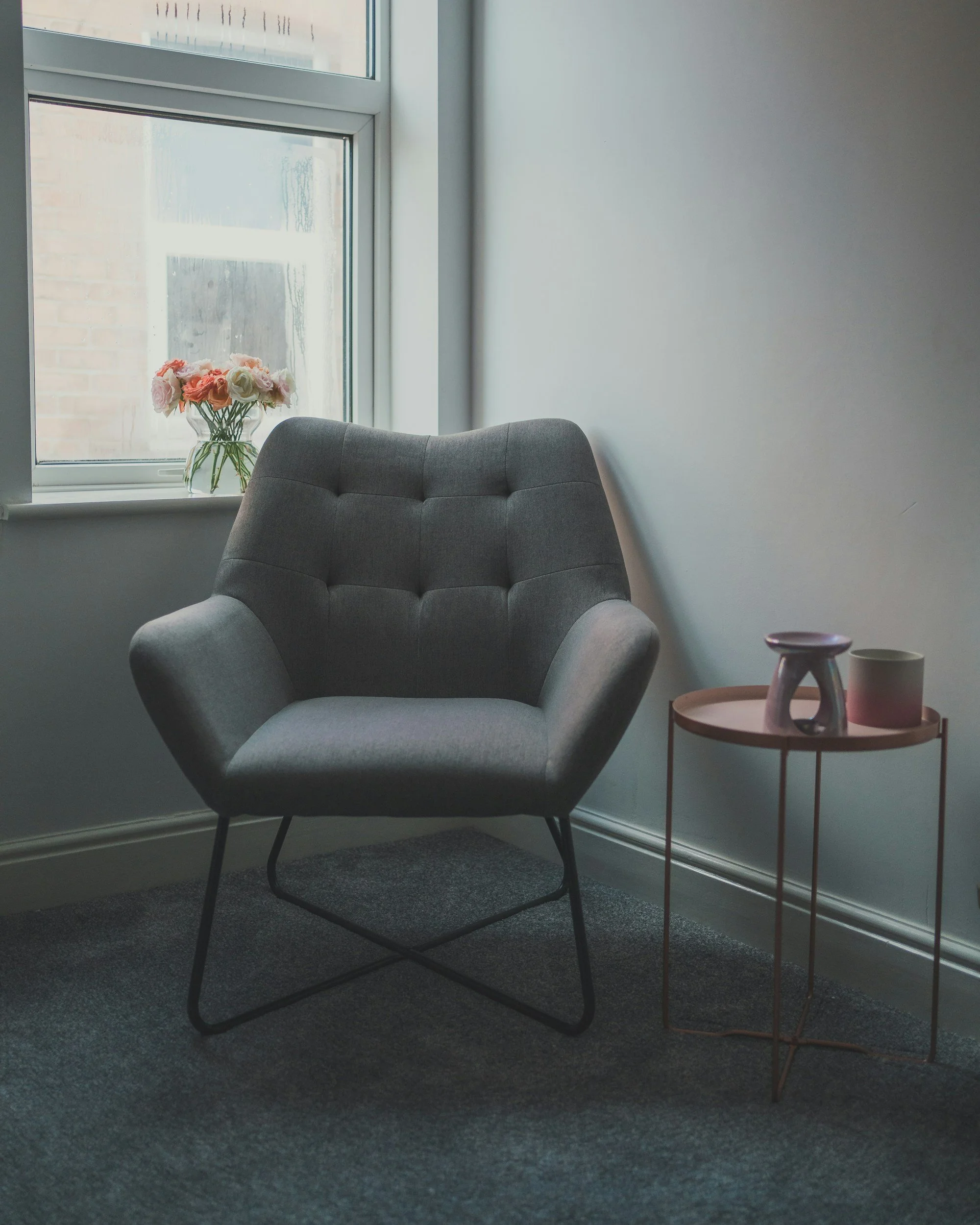 A cozy corner with a gray armchair, a small round side table with decorative vases, and a window with a flower arrangement on the sill.