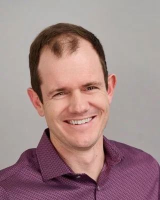 A man smiling with short dark hair, wearing a purple collared shirt, against a plain gray background.