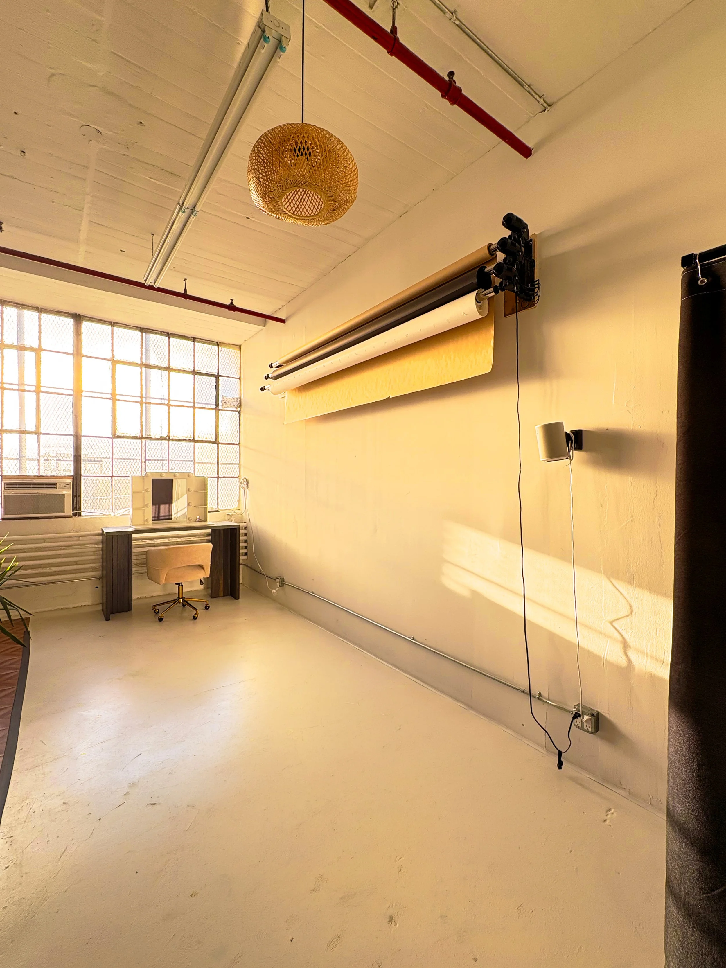 Empty room with large windowed wall, a ceiling lamp, a rolled-up backdrop, a small desk with a beige chair, and a black curtain.