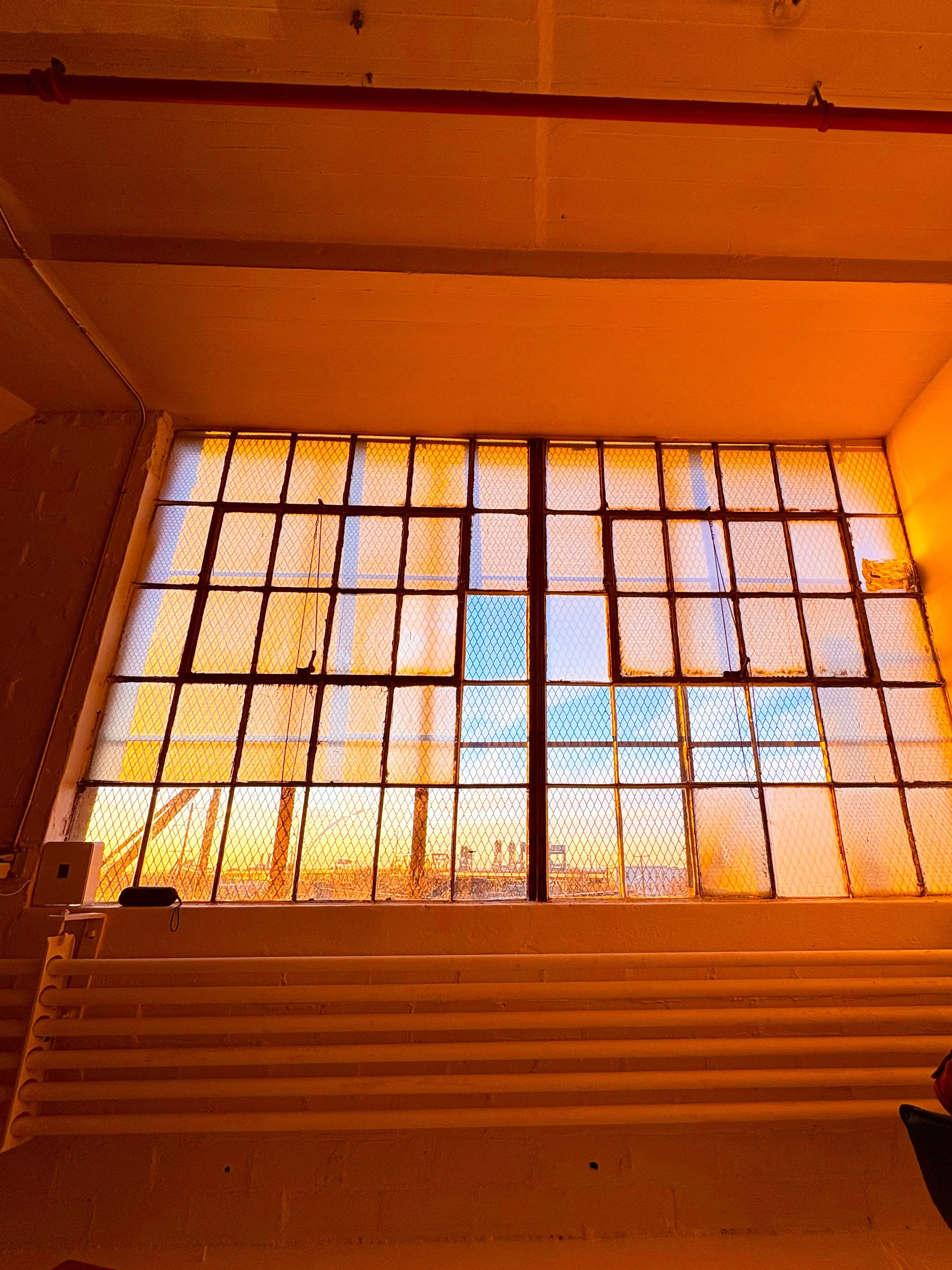 View of a large industrial-style window with metal grid frame, showing an orange sunset sky. There is a wooden slatted bench below the window and some objects on the wall on the left side.