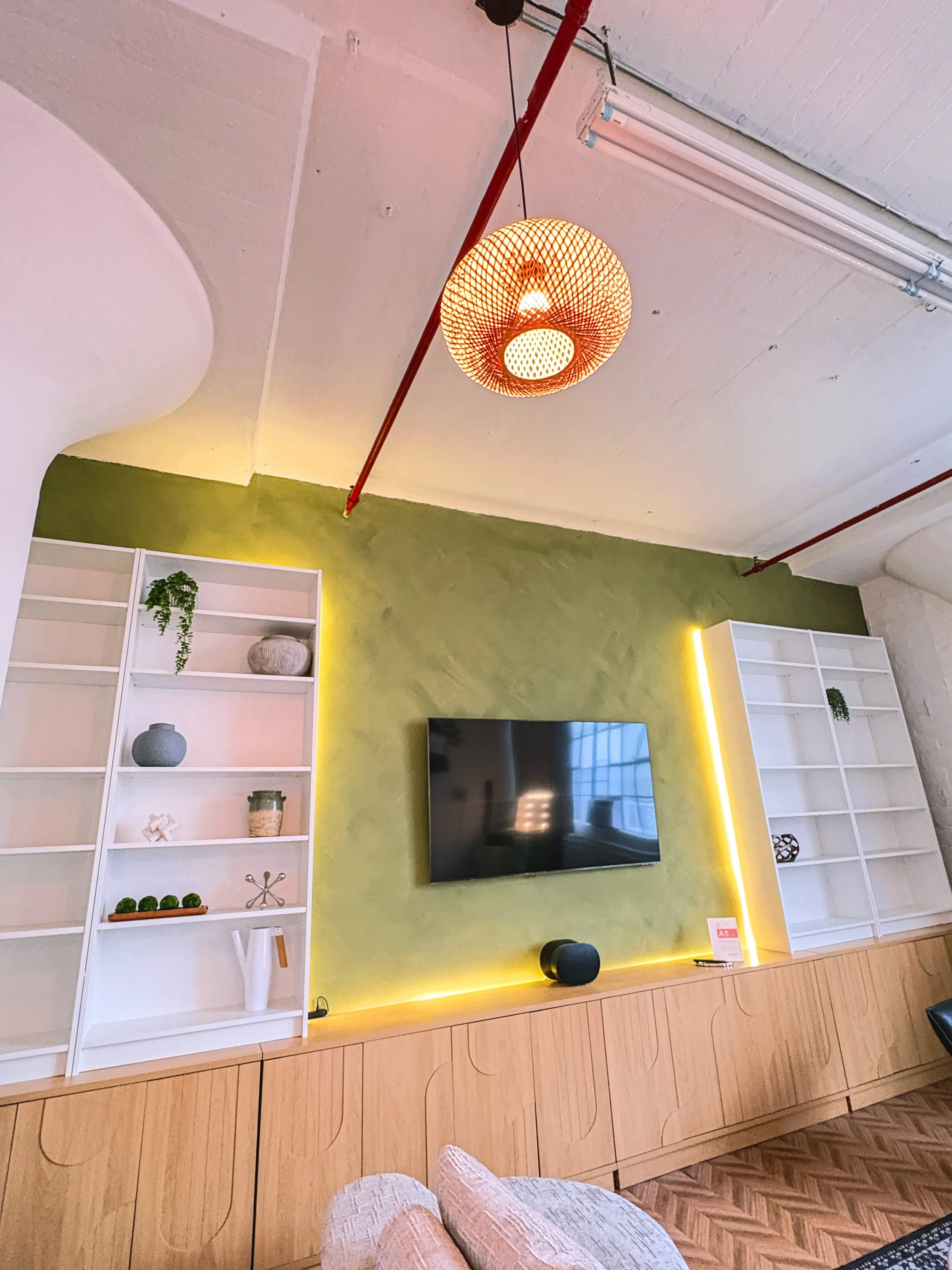 Living room with a wooden cabinet, a white bookshelf with decorative items, a green accent wall, a mounted TV, and a patterned floor, illuminated with yellow lighting.