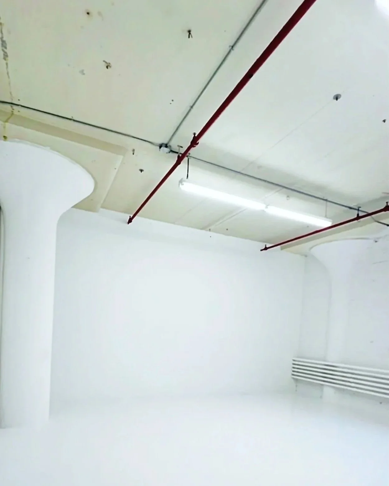 Empty room with white walls and ceiling, red piping running along the ceiling, and a white ventilation grille on the wall.