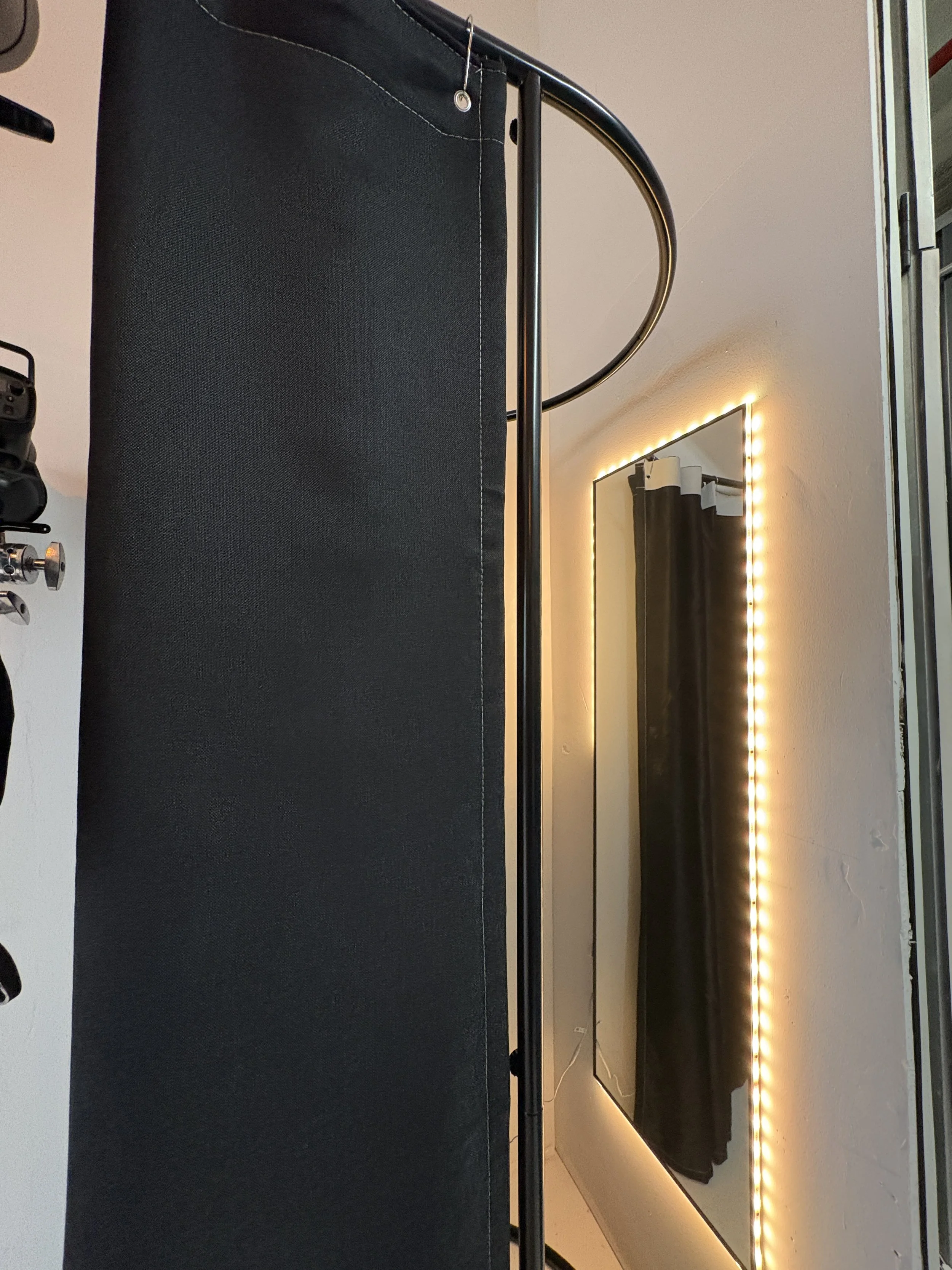 A black rectangular table with a black and silver pole on top, placed in front of a mirror with yellow LED strip lights around it. There is a black curtain behind the mirror and a white wall on the right side.