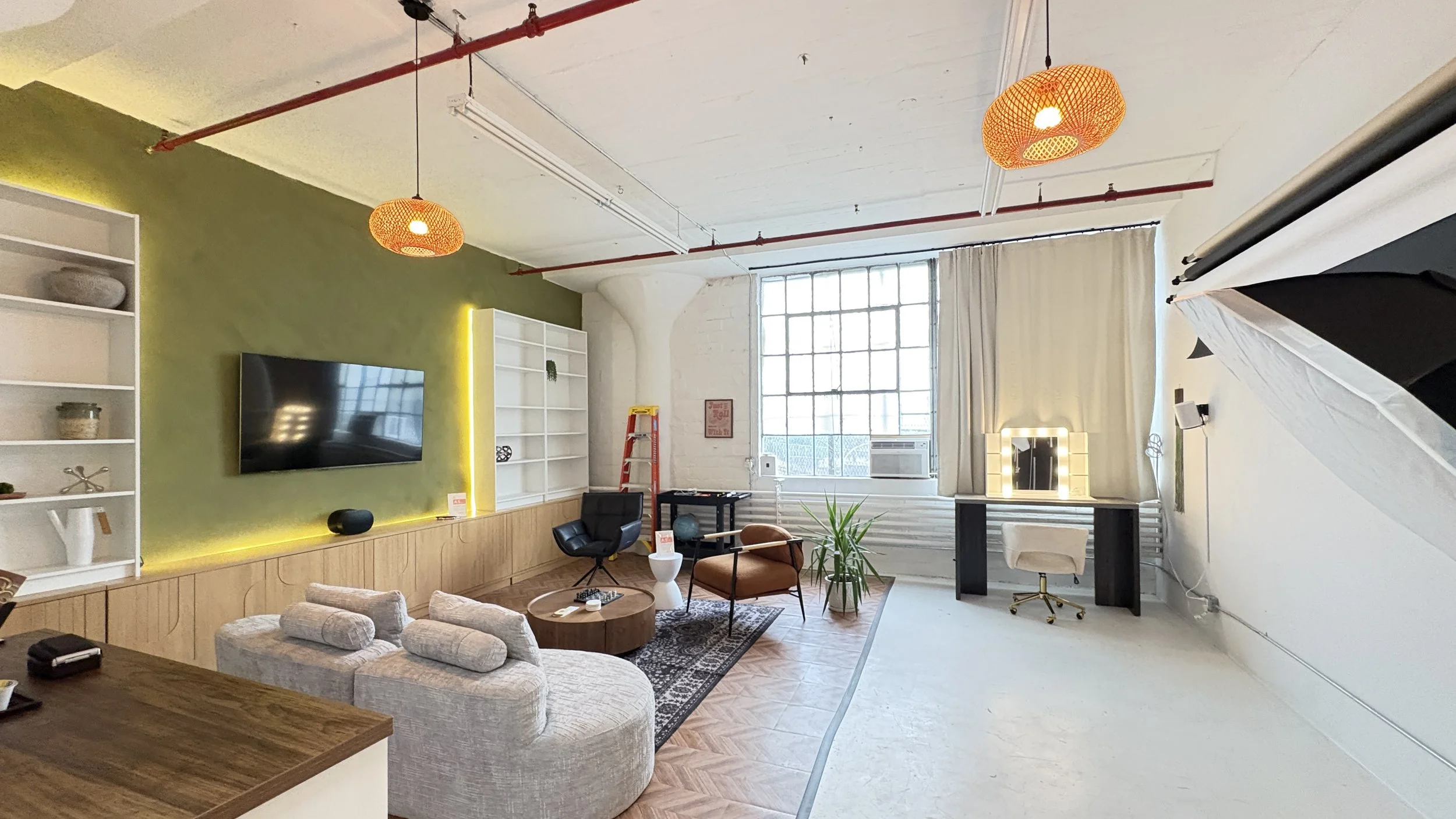 A modern, bright living room with a green accent wall, white shelves, a wall-mounted TV, and an exposed ceiling with red piping. There are orange pendant lights hanging from the ceiling, a window with white curtains, a black armchair, a wooden coffee table, a patterned rug, and a workspace with a desk, chair, and makeup mirror with lights. Decor includes plants and small decorative items.