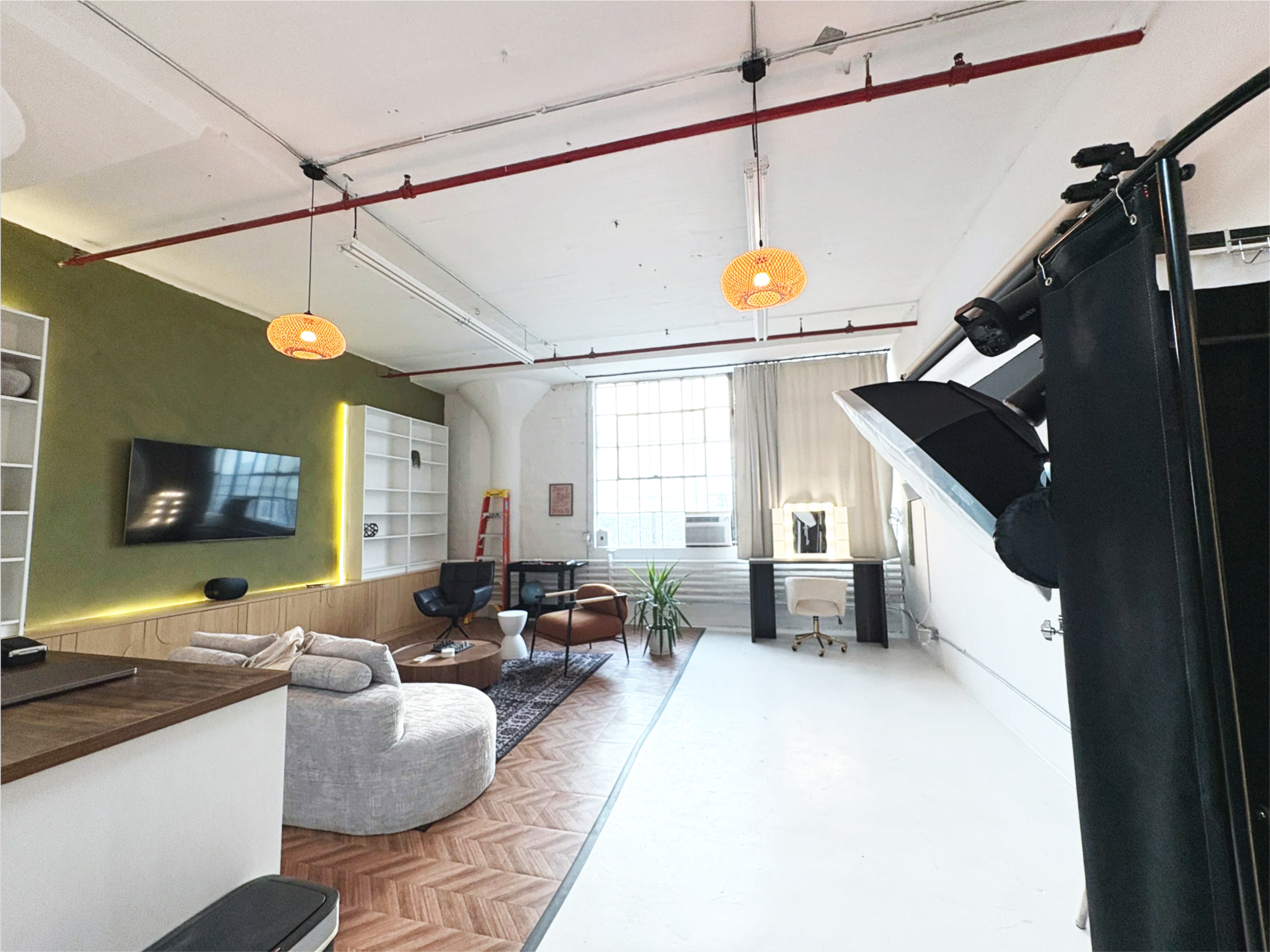 Interior of a modern loft-style apartment with an open space design, featuring a large flat-screen TV on a green accent wall, white shelving units, contemporary furniture including a cream-colored sectional sofa, black chairs, a rug, decorative plants, and large windows with curtains allowing natural light. Studio lighting fixtures and a photo shoot setup with equipment are also visible.