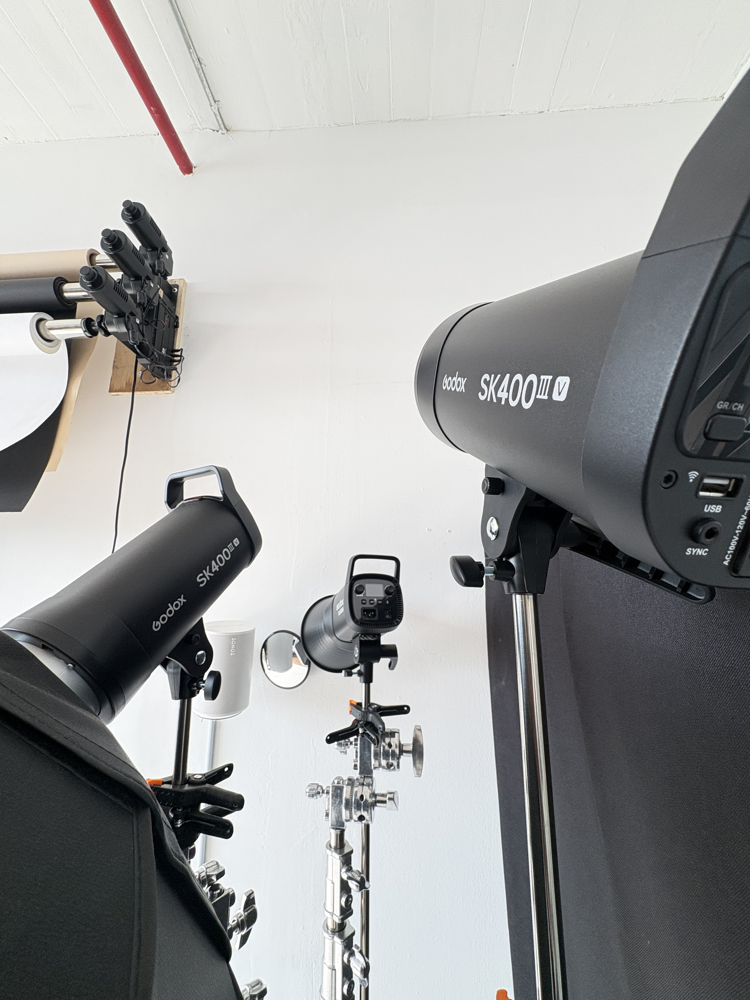 A photography studio setup with a Godox SK400II studio strobe light, a camera, and a light stand. The room has white walls and a concrete floor, with a red pipe running along the wall.