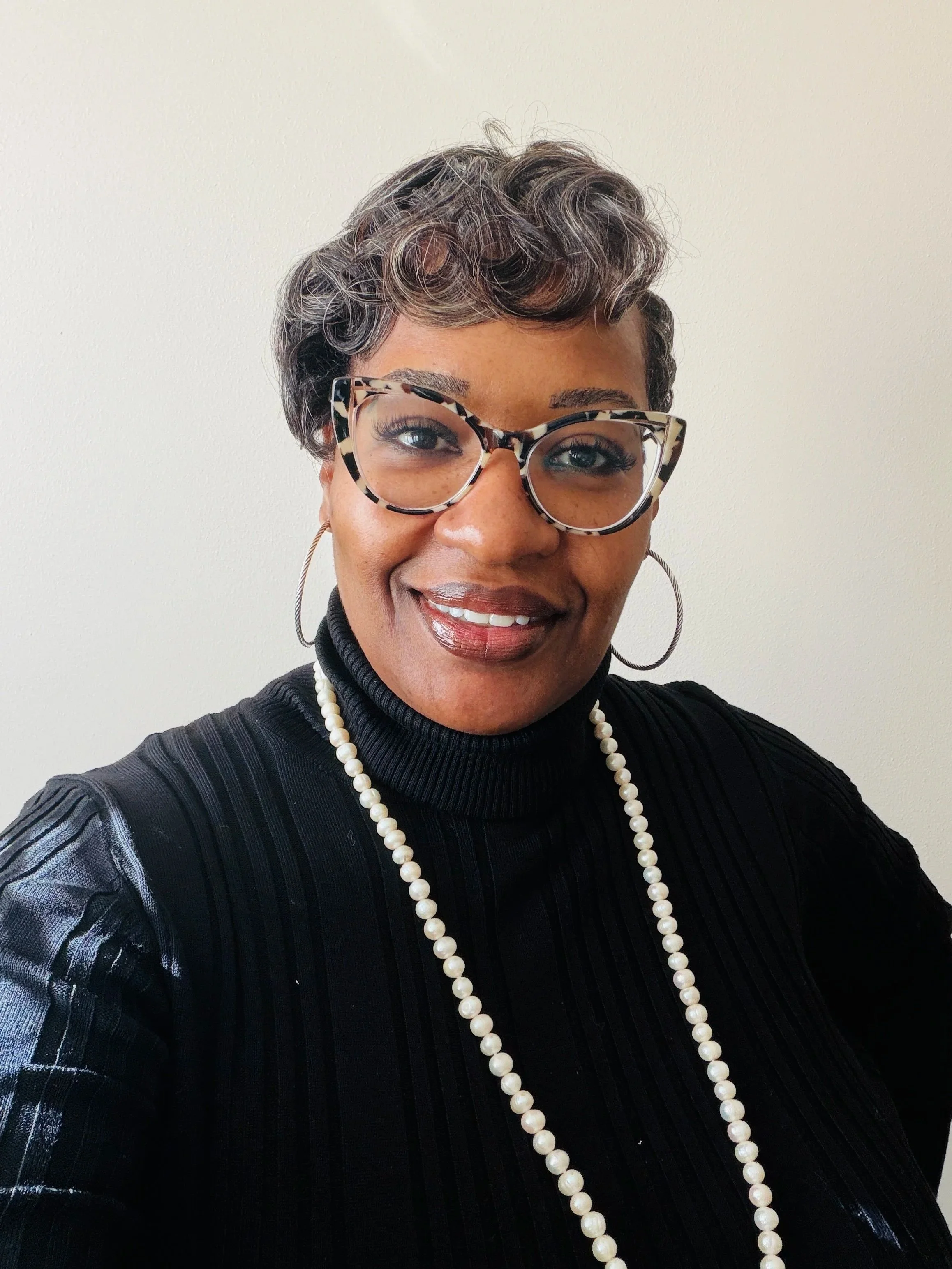 A woman wearing glasses, hoop earrings, a pearl necklace, and a black turtleneck sweater with textured sleeves, smiling against a plain light-colored background.