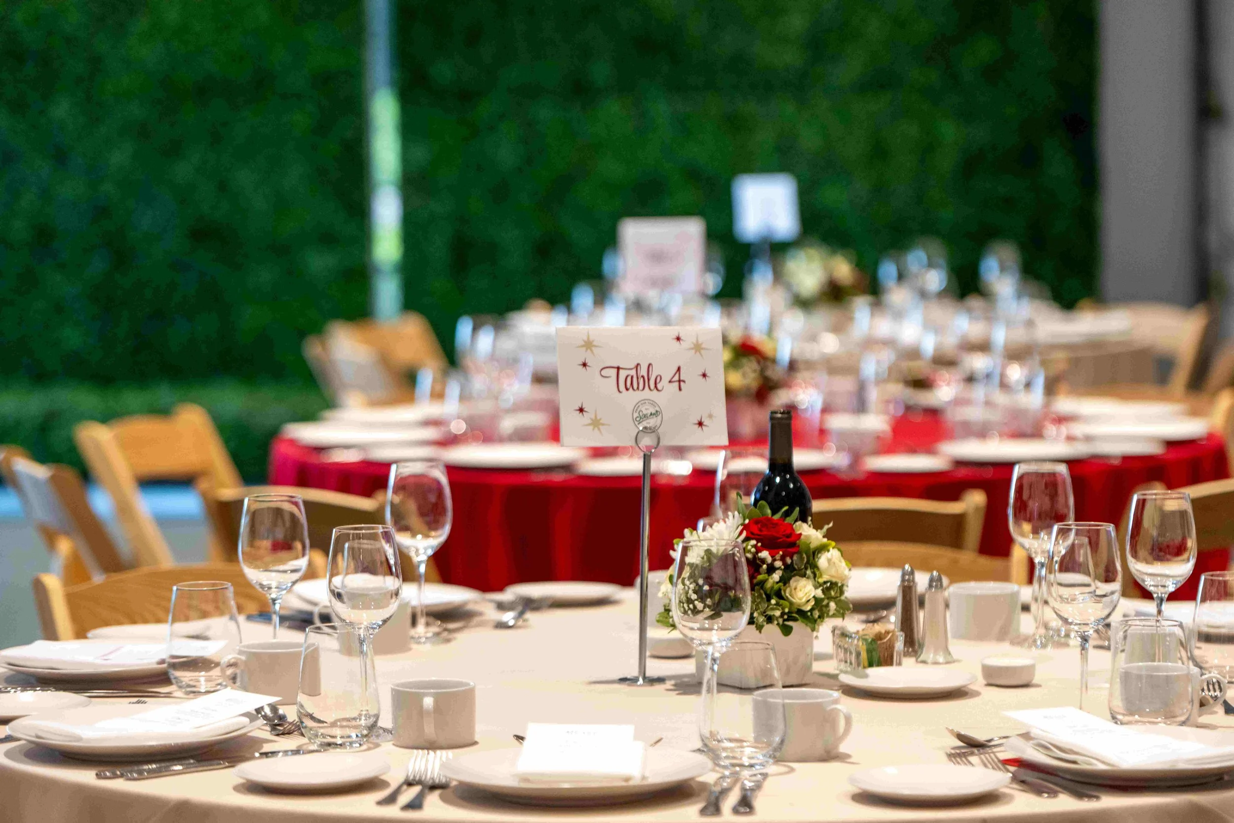 A banquet table set for a meal, with wine glasses, plates, and silverware, decorated with a floral centerpiece and a sign indicating it is for 'Table 4' in a room with green walls and additional tables.