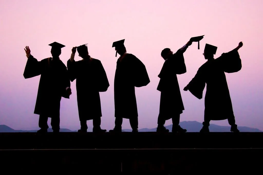 Silhouettes of five people wearing graduation gowns and caps, celebrating graduation against a pink and purple sky backdrop.