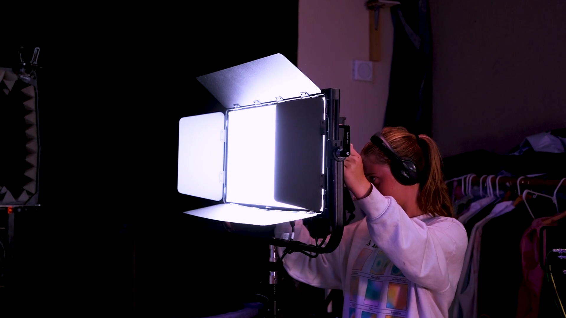 A woman operating professional photography or video equipment with a large LED light panel, in a dimly lit room with a closet in the background.