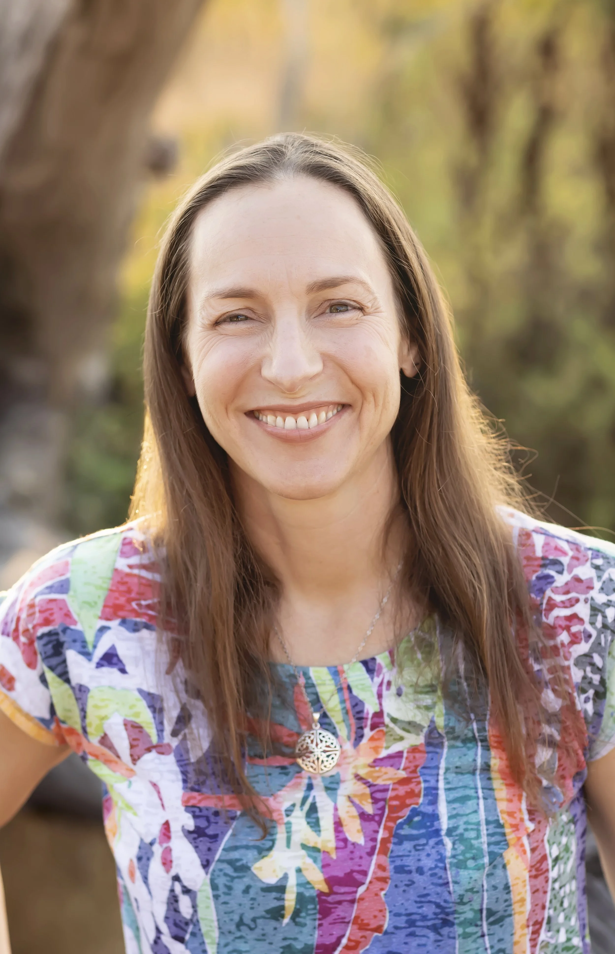 A woman with long reddish-brown hair smiling outdoors in a colorful, patterned top, wearing a silver pendant necklace with a tree design.
