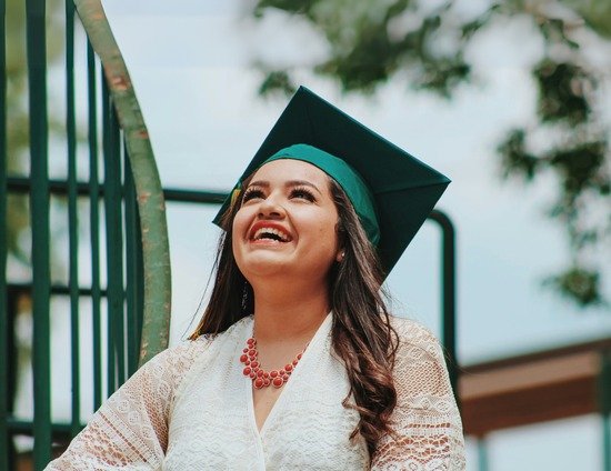 A woman wearing a graduation cap and gown, smiling and looking upwards outdoors.