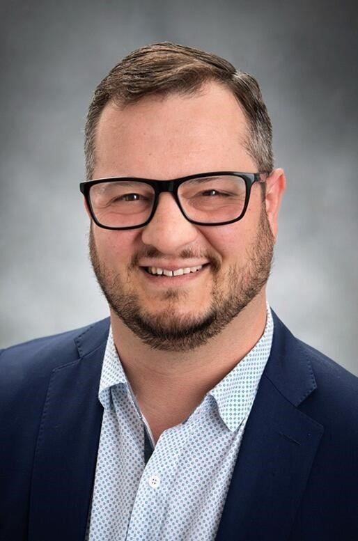 A man with short hair, beard, and glasses smiling, wearing a navy blazer and a light blue dress shirt with small polka dots, against a neutral background.