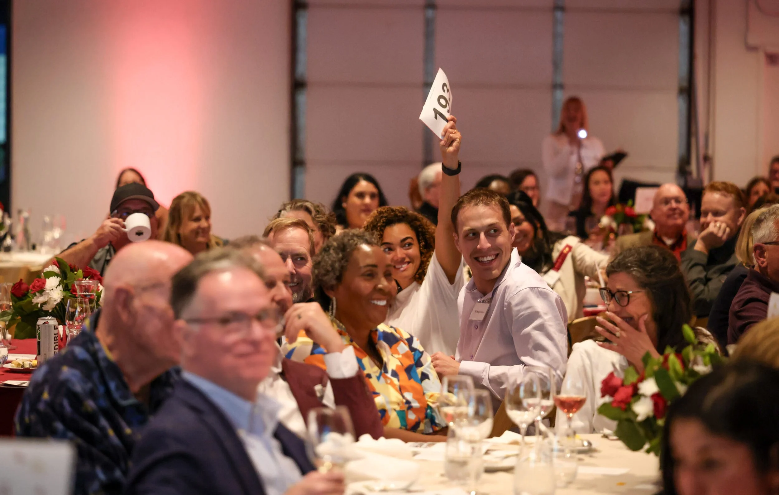 A group of people sitting at tables during a formal event, smiling and engaging, with one person holding up a sign with the number 109, in a decorated banquet hall.