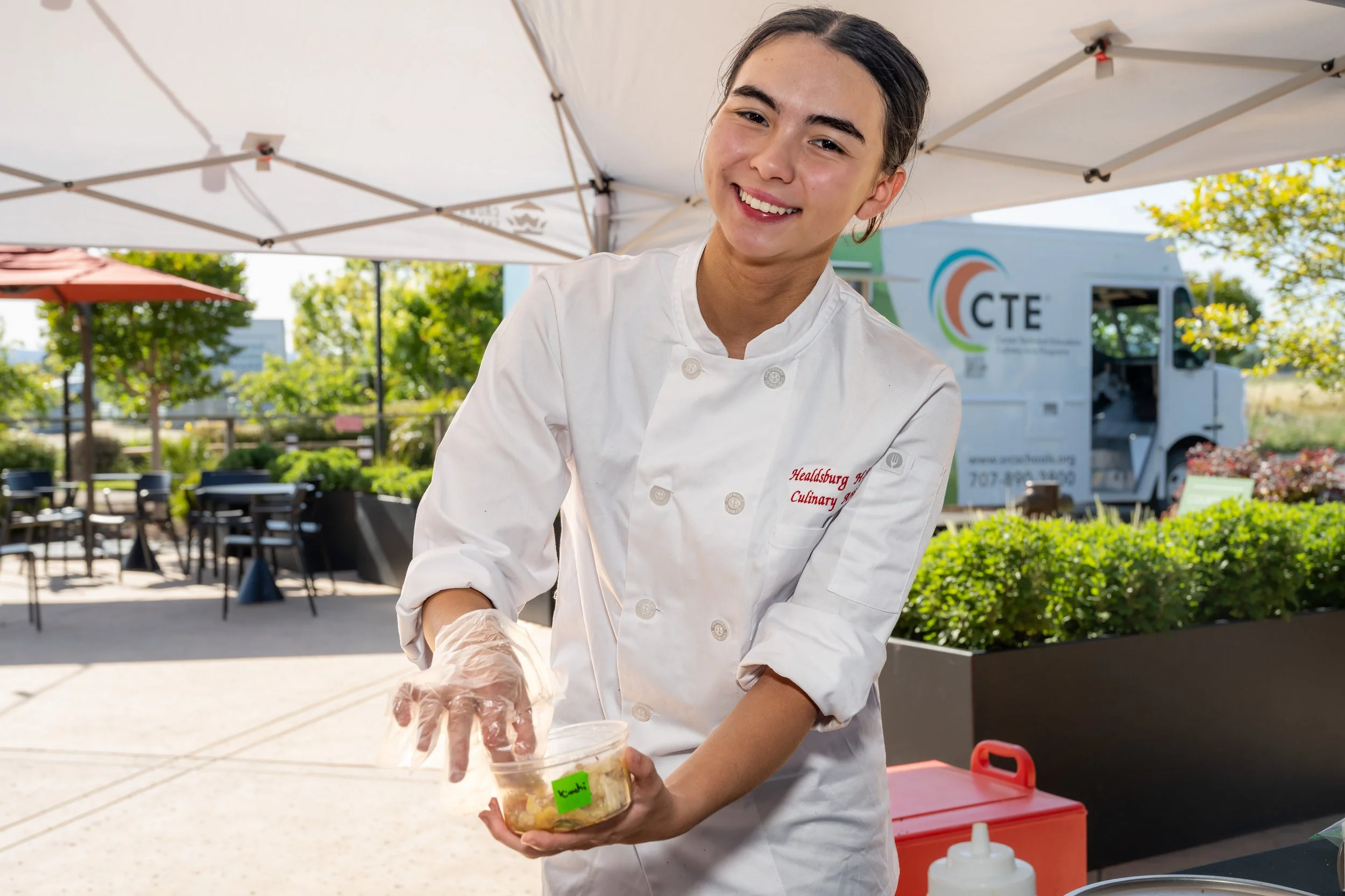 A young woman in a white chef's coat is smiling and preparing food outdoors under a white canopy. She is wearing plastic gloves and holding a clear plastic container with food inside. There are tables, chairs, and a food truck with a colorful logo in the background.