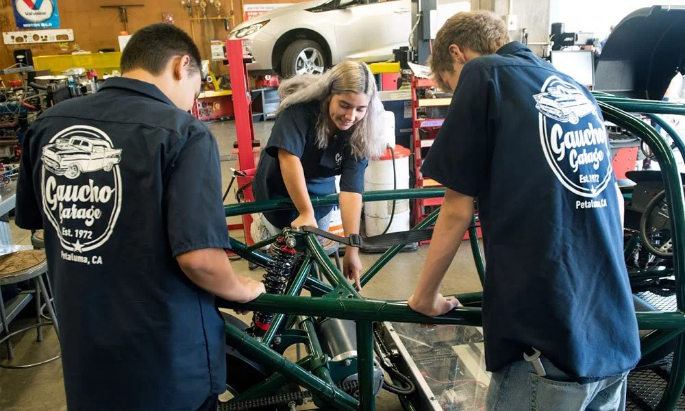 Three people working on a green vehicle frame in a garage workshop. Two are wearing navy shirts with the 'Gaucho Garage' logo on the back. The woman in the middle has long gray hair and is smiling while working on the frame.