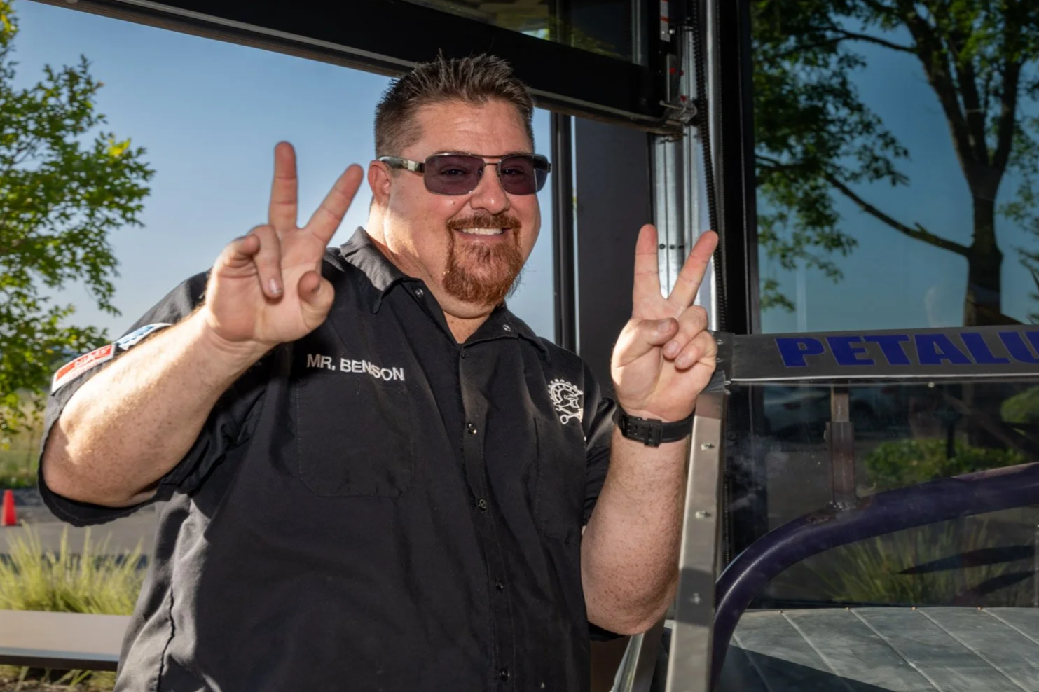 A man with a beard, sunglasses, and a black uniform smiling and making peace signs with both hands. The uniform has a patch on the shoulder and the name tag 'MR. BENSON'. He is standing outside near a vehicle with a sign that reads 'PETALU' and some trees are visible in the background.