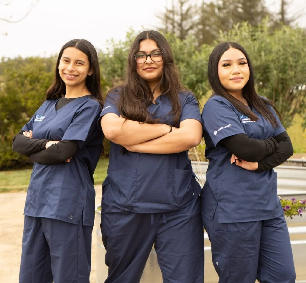 Three women dressed in navy medical scrubs standing outdoors with arms crossed, smiling confidently.