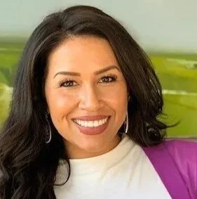 Close-up of a smiling woman with long dark hair, wearing a white top and purple blazer, against a blurred green background.