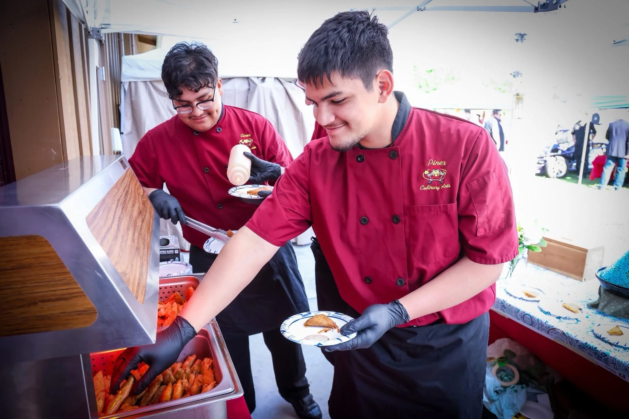 Two male chefs, wearing red uniforms with name tags that read 'Piner Culinary Arts,' serve food at an outdoor event. They are using black gloves, with one holding a plate and serving from a tray of fried food, possibly chicken wings, under a large white canopy. In the background, there are people, tables, and a few parked vehicles.