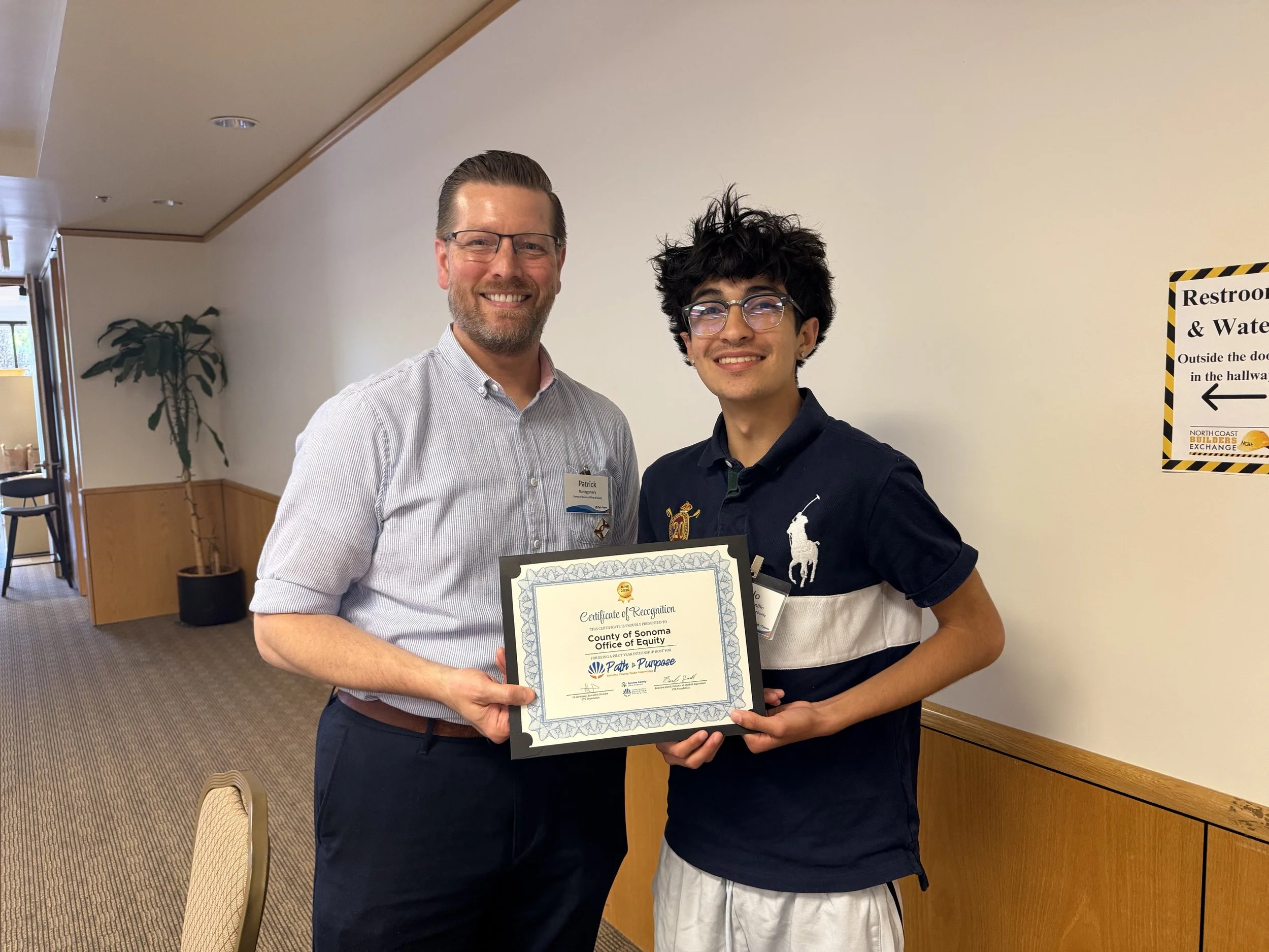 Two men standing together indoors, smiling, one holding a certificate. The man on the left has glasses, a beard, and a striped shirt; the younger man on the right has curly hair, glasses, and a dark polo shirt with a logo.