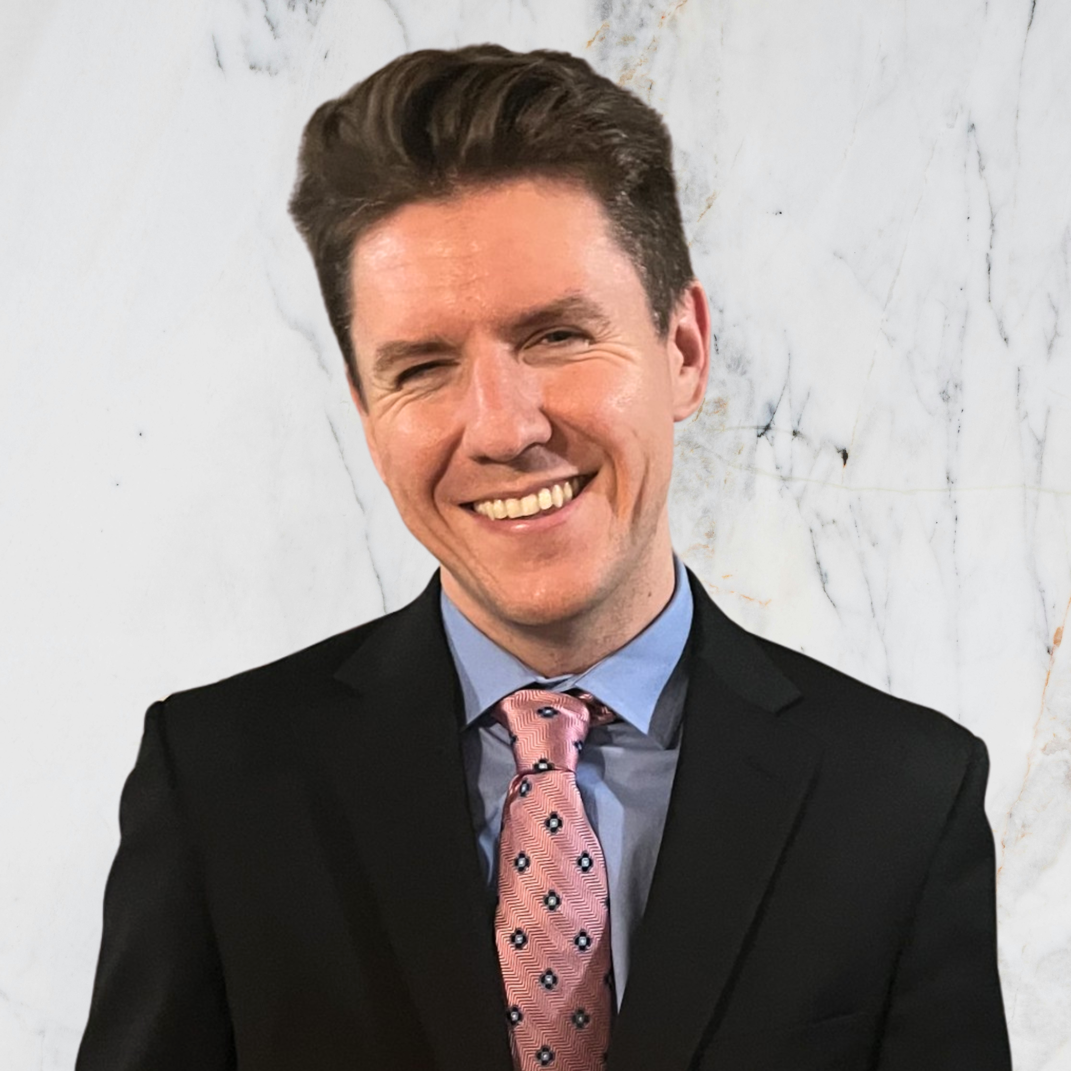Smiling man wearing a black suit, light blue shirt, and pink tie with black dots, standing against a white marble background.