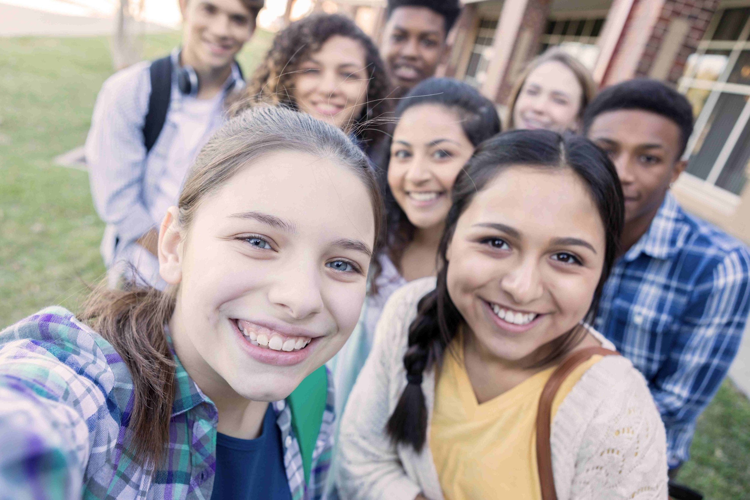 Group of diverse young students smiling outdoors on a school campus, taking a selfie together.