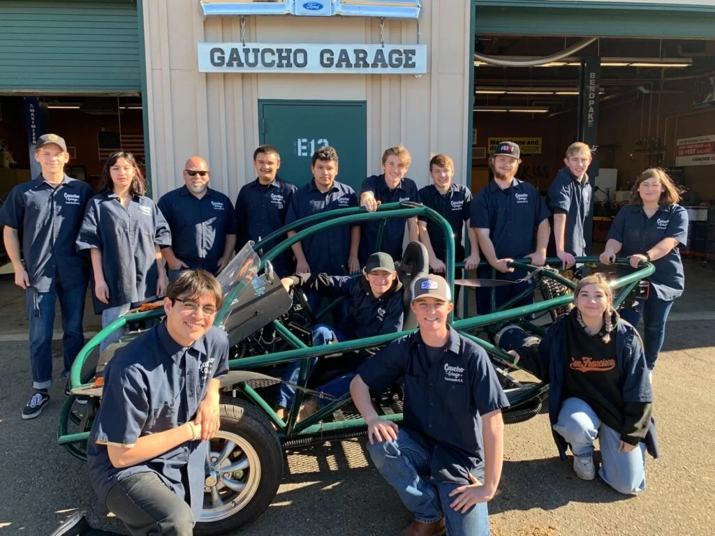 Group of people, including students and a few adults, standing and kneeling in front of a greensand automotive project vehicle outside a garage called Gaucho Garage.