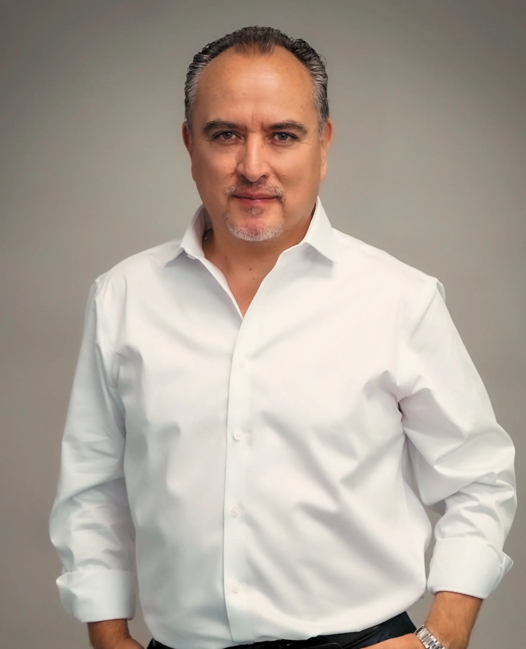 A man wearing a white dress shirt with the top button undone, standing against a plain gray background.