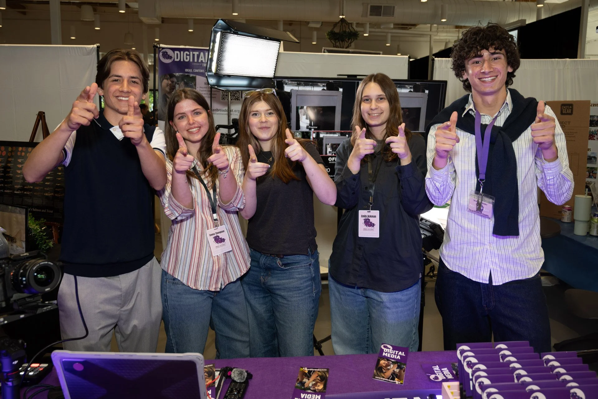 Five young people standing together at a booth, smiling and pointing at the camera, with equipment and promotional materials on the table in front of them, at an indoor event or conference.