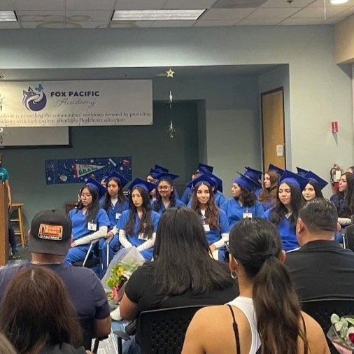Group of high school graduates in blue caps and gowns seated on stage at a graduation ceremony, with audience members watching.