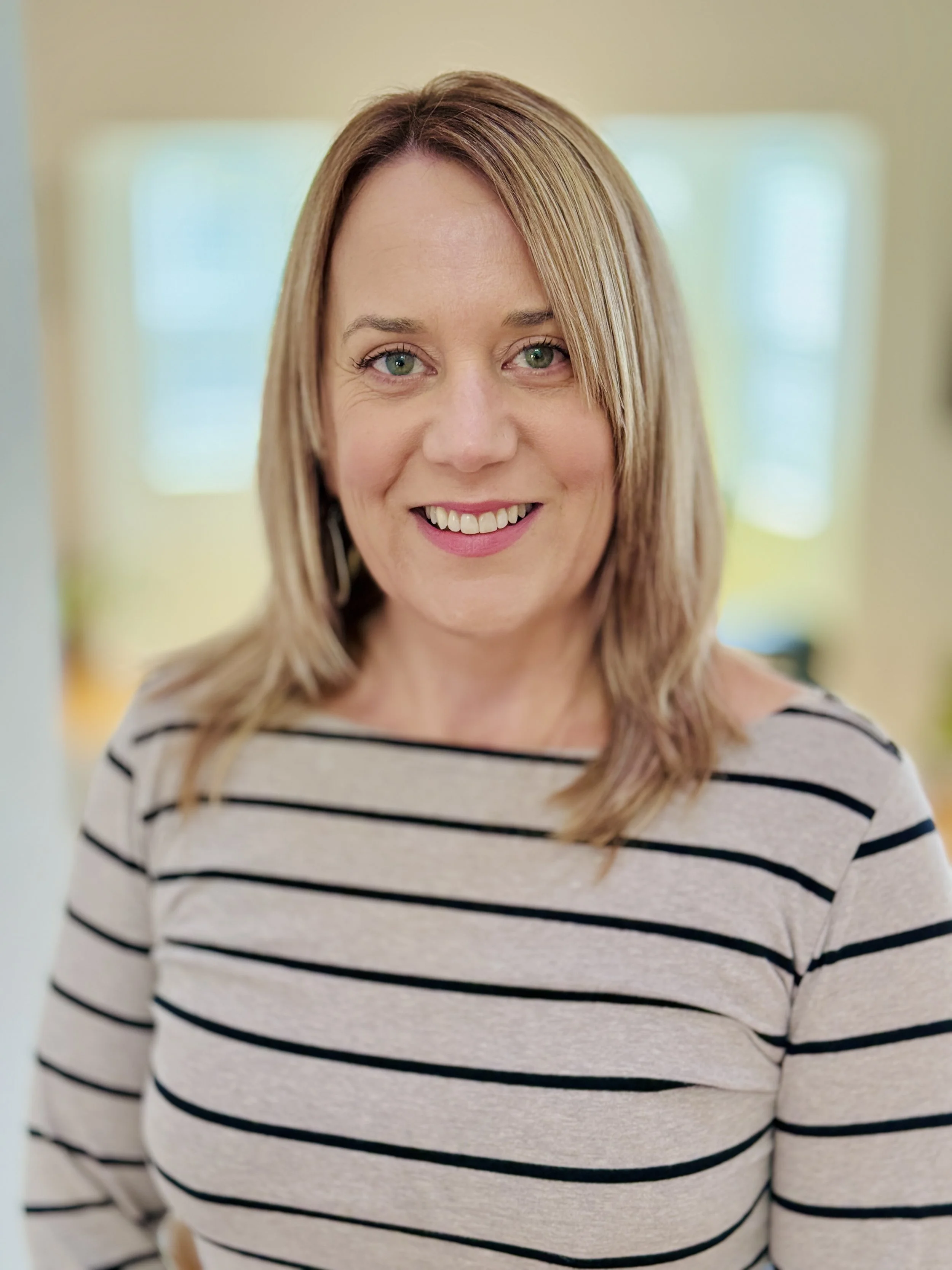 Close-up of a smiling woman with shoulder-length blonde hair wearing a beige and black striped shirt, standing indoors with blurred windows in the background.