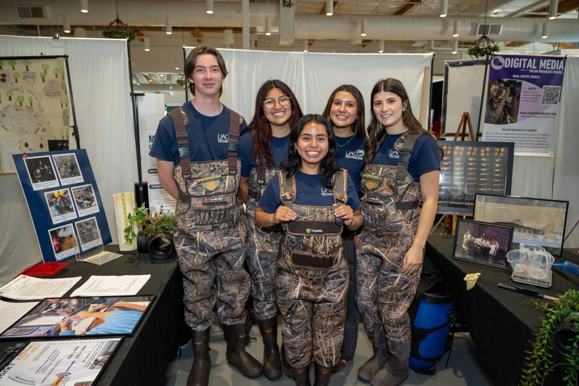 Group of five young adults at an indoor event, wearing camouflage pants and navy shirts with social media icons, standing behind a table with informational displays and brochures.