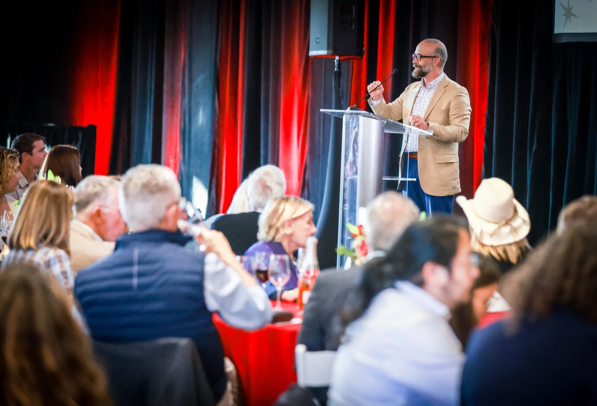 A man with glasses and a beard, wearing a beige blazer and checkered shirt, stands at a clear podium speaking into a microphone at a conference or event. The background features red curtains, and the audience is seated at round tables.