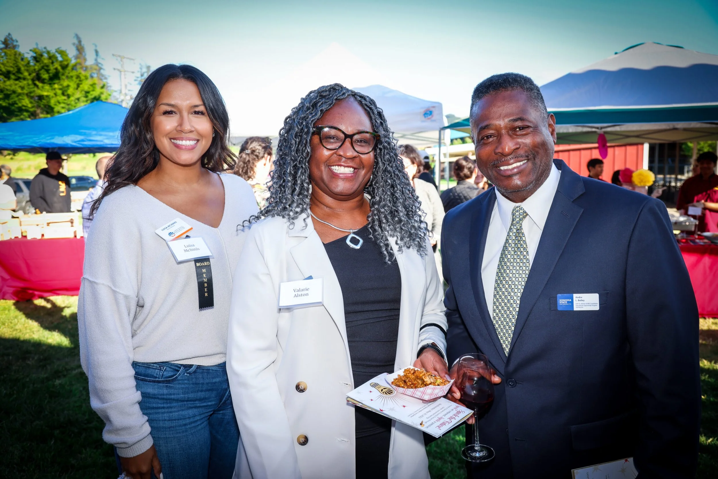 Three people standing outdoors at a daytime event with tents behind them. The woman on the left has dark hair and is wearing a white sweater. The woman in the middle has curly gray hair, glasses, and is wearing a white blazer. The man on the right has short gray hair, is in a dark blue suit, and is holding a glass of red wine. They are smiling and looking at the camera.