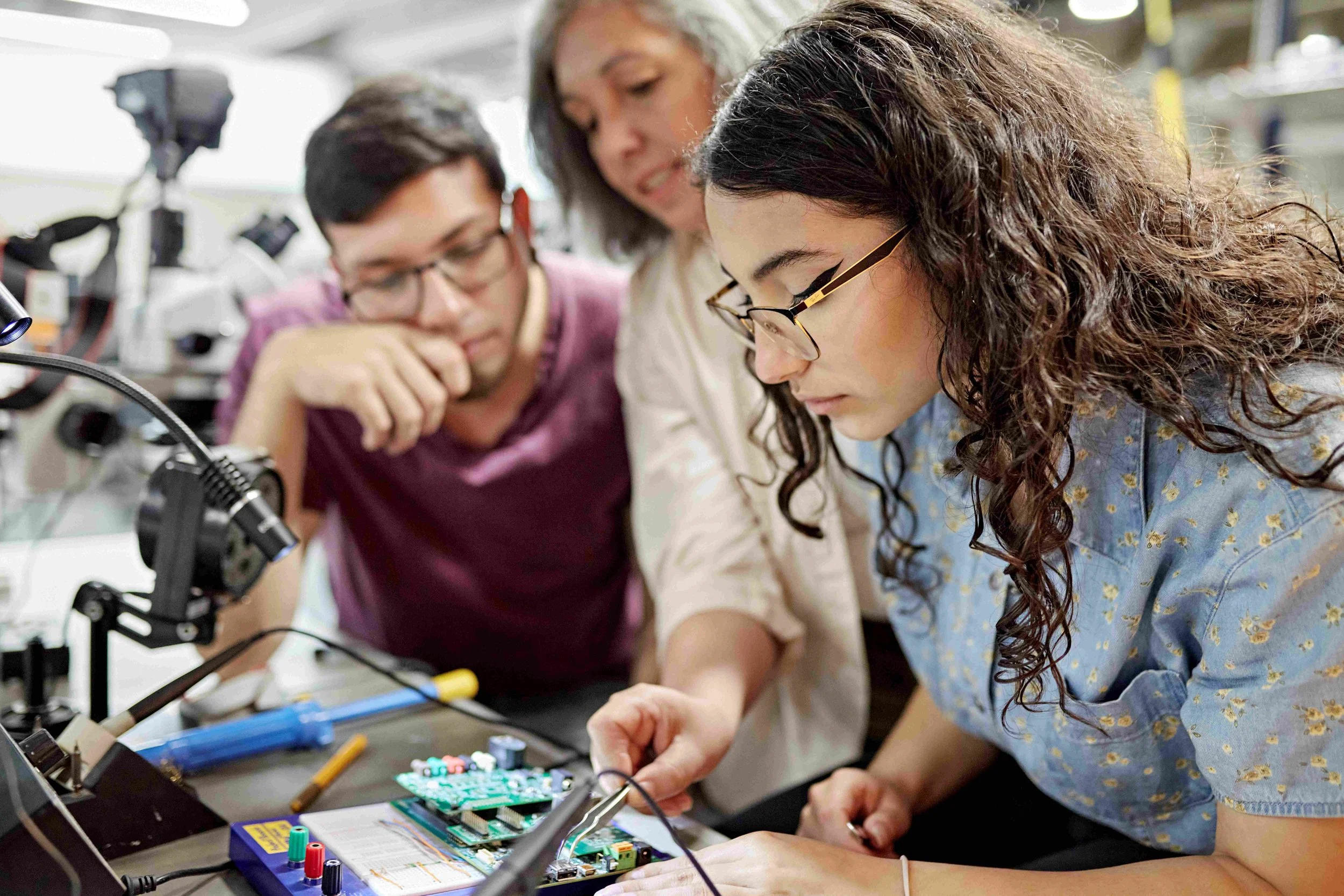 Group of three young women working together on an electronics project with a circuit board and tools on a laboratory table.