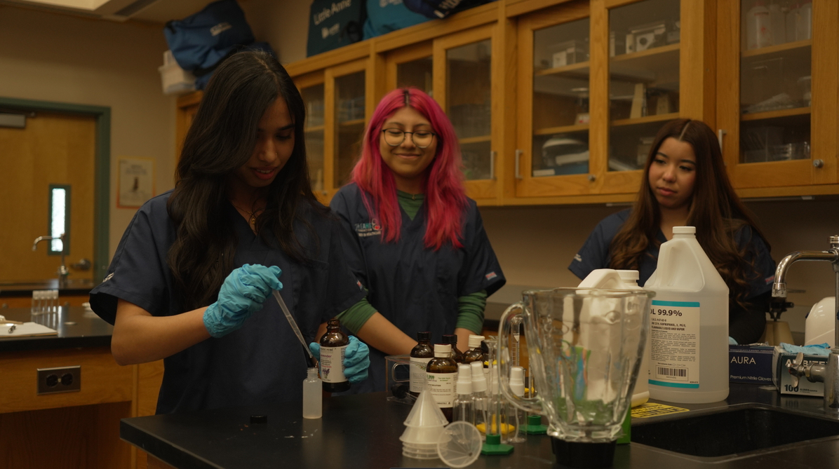 Three young women in a science laboratory. One is using a pipette to transfer liquid into a test tube, while the other two watch. The workspace has bottles, measuring devices, and lab equipment.