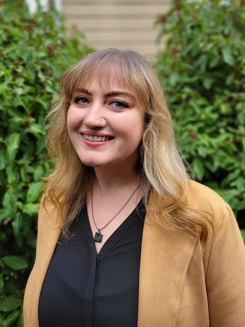 A young woman with blonde hair, wearing a black top and a tan jacket, smiling outdoors with green plants in the background.