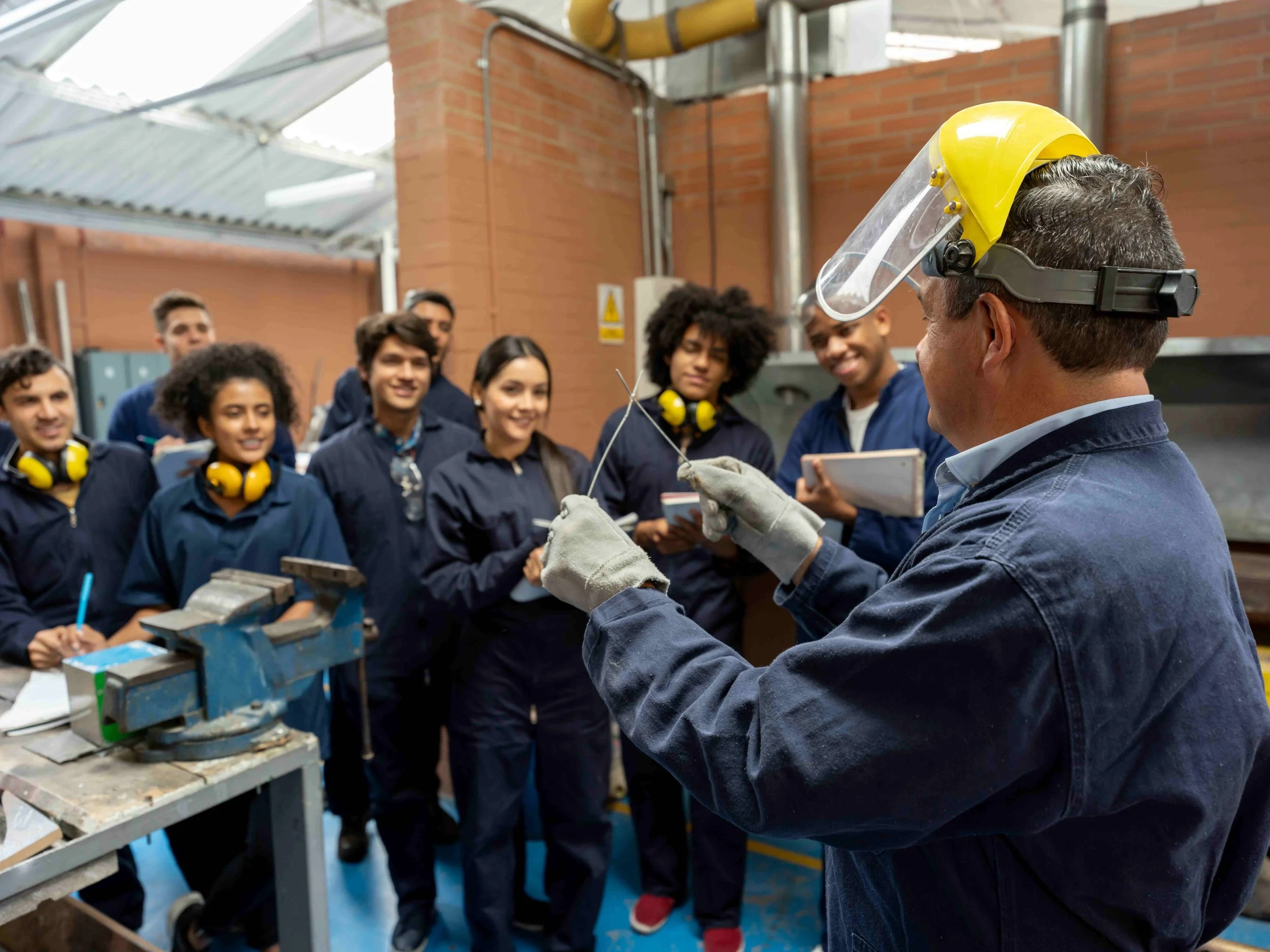 A man wearing a yellow hard hat and safety gloves shows a group of young workers in safety gear, listening attentively in an industrial workshop with brick walls and metal pipes.
