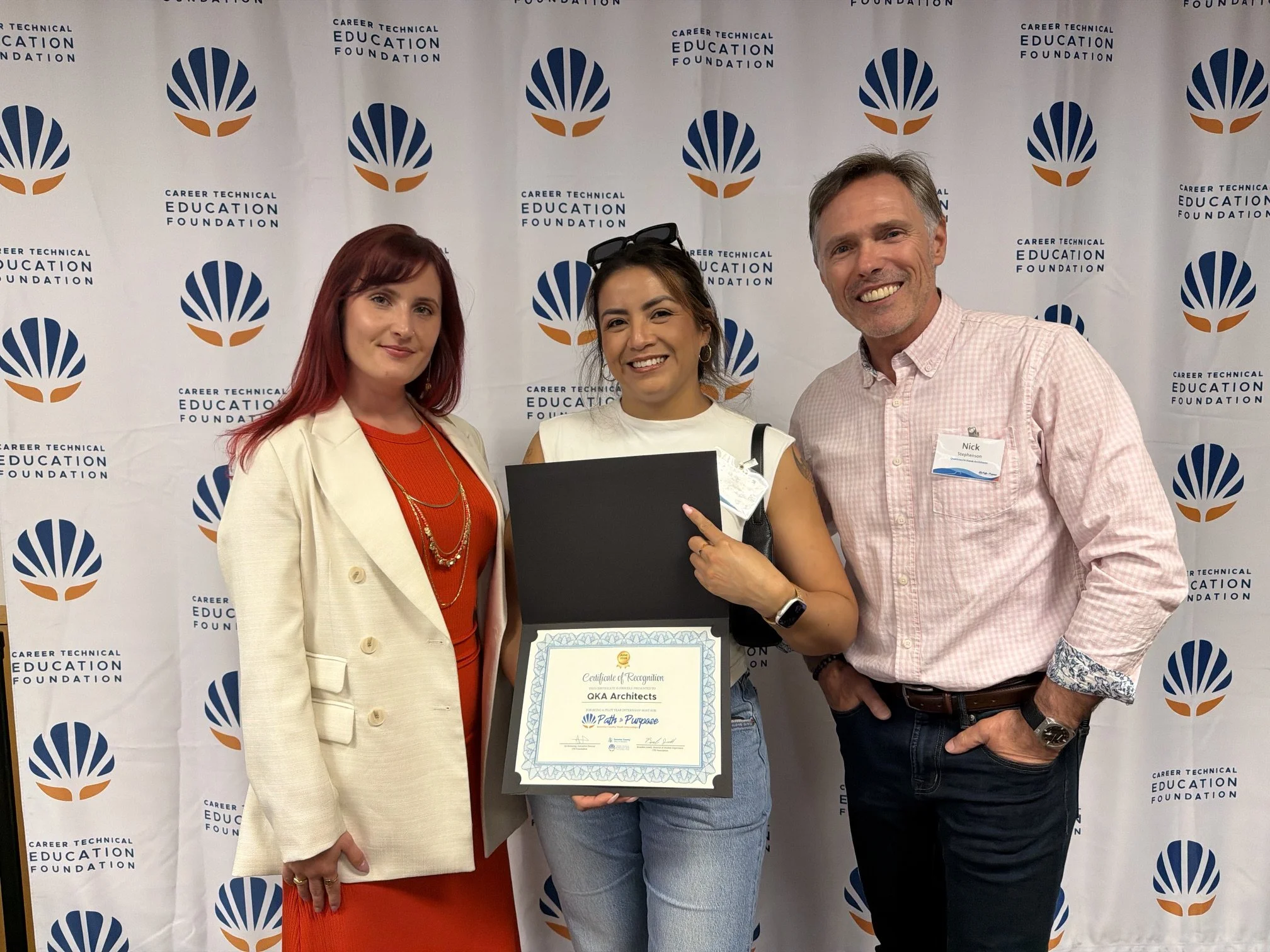 Three people standing in front of a backdrop with a logo and text. The person in the middle is holding a framed certificate. The person on the right is wearing a name tag that reads "Nick." The backdrop displays the logo and the words "Career Technical Education Foundation." The woman on the left has red hair, and the woman in the middle has dark hair and is wearing glasses on her head.