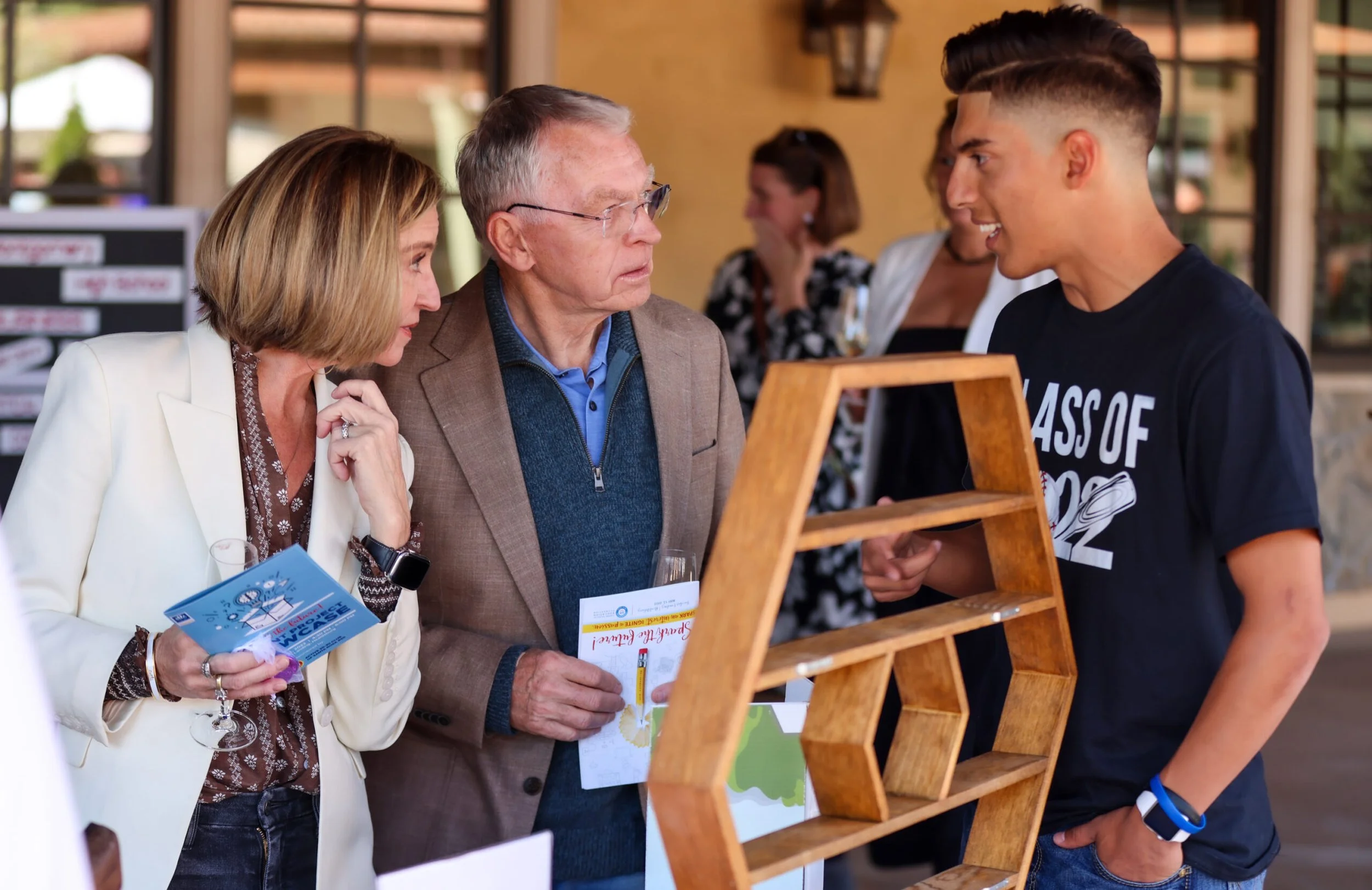 Three people engaged in conversation at an outdoor event, with one person holding brochures and another presenting a wooden display stand.
