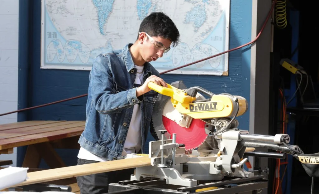 A young man wearing safety goggles and a denim jacket operating a yellow DEWALT miter saw to cut a piece of wood in a workshop with a world map on the wall.
