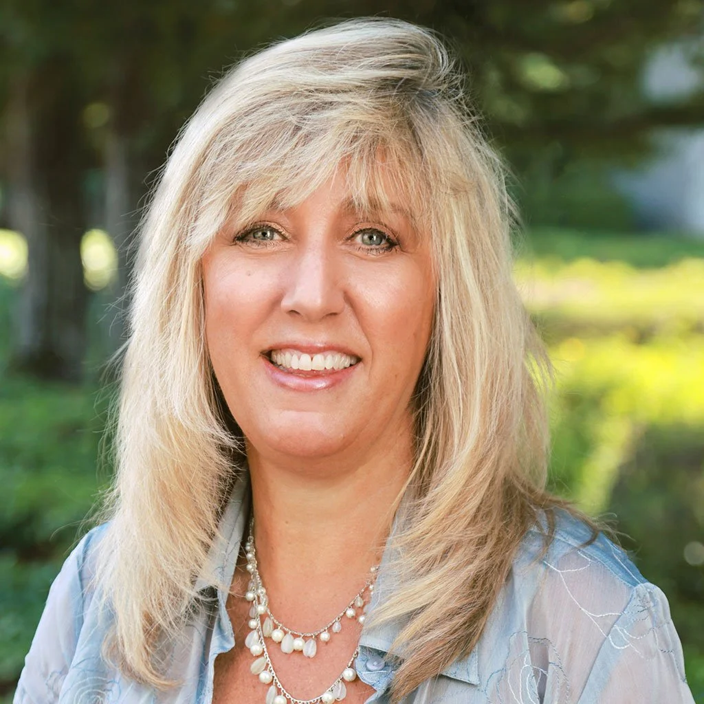 A smiling middle-aged woman with blonde hair and blue eyes, wearing a pearl necklace and a light-colored blouse, outdoors with a blurred green background.