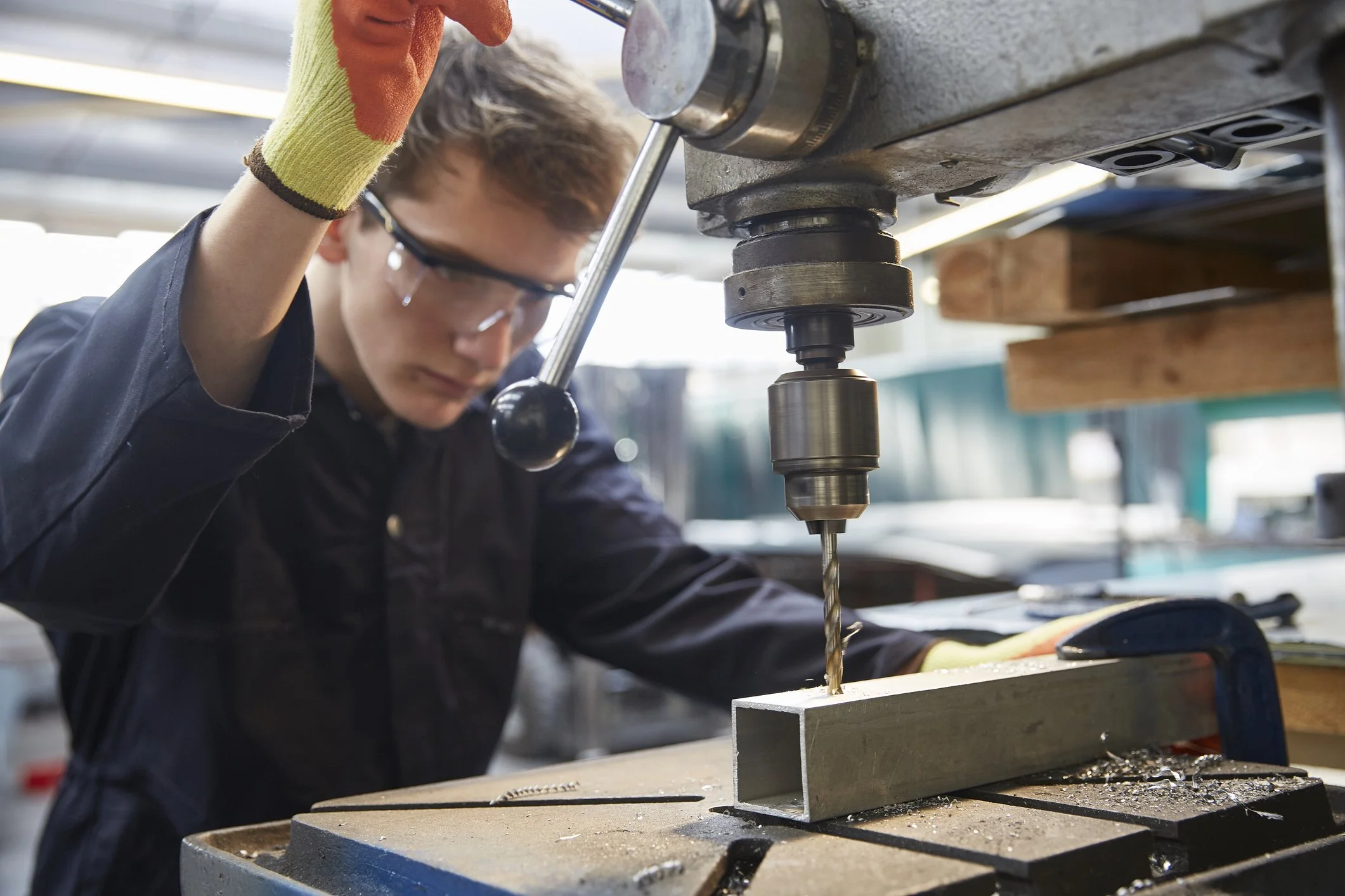 A person wearing safety glasses and gloves working with a drill press on a metal piece in a workshop.