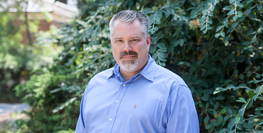 A middle-aged man with gray hair and a beard standing outdoors in front of green bushes, wearing a light blue dress shirt.